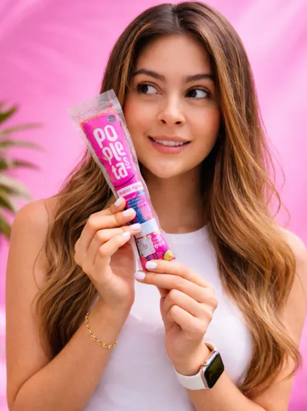 Mujer sonriente sostiene una paleta Popletas en su empaque rosado frente a un fondo rosa